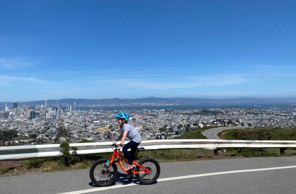 a young cyclist on a red bike riding on a hillside road with the city in the background