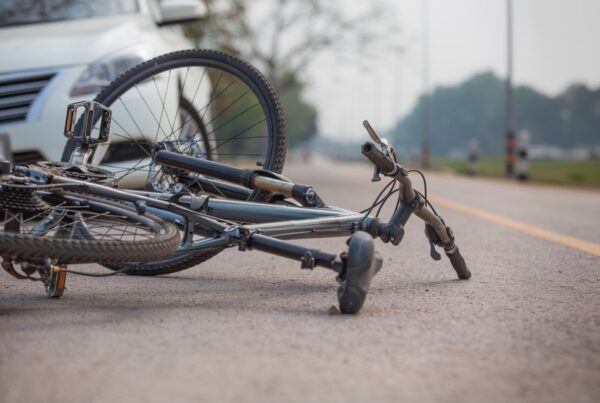 A bike lies on its side on a road next to a white car, suggesting a recent accident. The scene is outdoors with trees in the background.